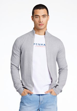 Ljusgrå zip-up cardigan med ribbat mönster, bärs över en vit t-shirt med blå och röda texter. Modellen står med händerna i fickorna.