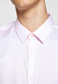 Jasnoróżowa koszula zapinana na guziki o fakturowanej tkaninie, z klasycznym kołnierzykiem i listwą guzikową z okrągłymi, białymi guzikami.