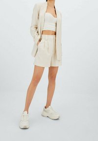 Blazer en lin beige associé à un crop top blanc froncé et un short beige taillé. Le modèle porte des baskets blanches. Design simple et épuré.