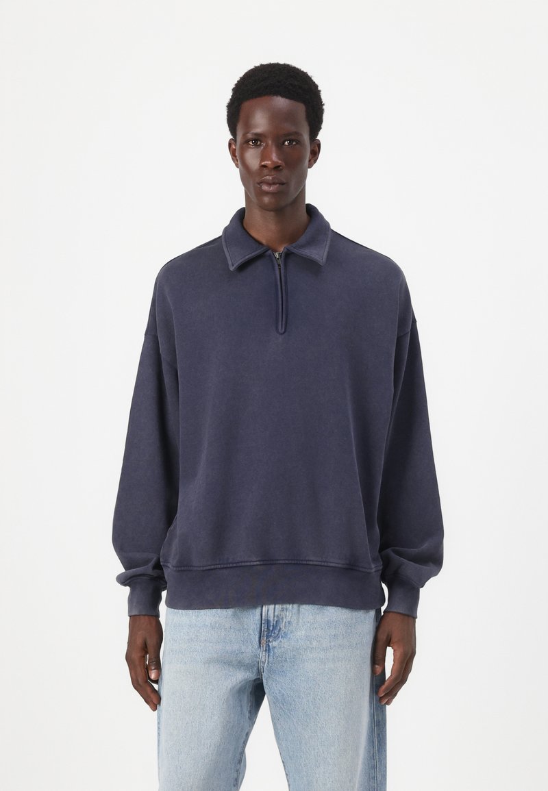 Felpa blu navy con zip a metà, realizzata in tessuto morbido, con colletto e maniche a polsini. Texture vissuta e vestibilità comoda.