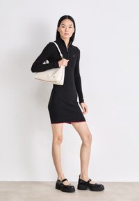 Rochie neagră cu dungi, cu accente roșii, mâneci lungi și guler cu fermoar. Modelul ține o geantă albă de umăr cu detalii aurii și poartă pantofi negri masivi.