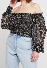 Black off-the-shoulder top with floral print, smocked bodice, and sheer, puffed sleeves. Paired with high-waisted light denim jeans.