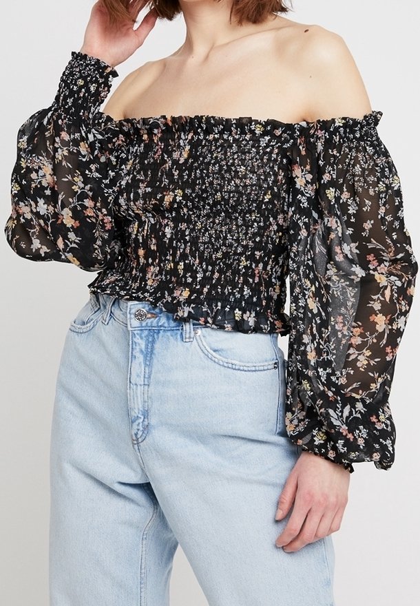 Black off-the-shoulder top with floral print, smocked bodice, and sheer, puffed sleeves. Paired with high-waisted light denim jeans.