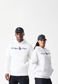 Os hoodies brancos apresentam "Los Angeles Dodgers" e "LA" em letras navy, combinados com bonés pretos. Estilo casual, corte relaxado com bolsos frontais.