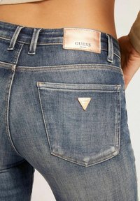 Jeans de mezclilla en un lavado azul desgastado, con un logo triangular de Guess en el bolsillo trasero y un parche de cuero texturizado en la cinturilla.