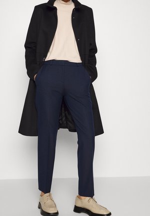 Personne portant un manteau noir, un col roulé beige, un pantalon bleu marine sur mesure et des chaussures beiges à lacets, debout les mains dans les poches.