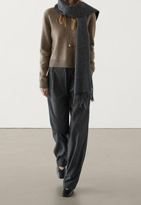 Maglione marrone in lana con orlo a coste, sciarpa grigia con frange, pantaloni grigi a righe sottili e scarpe basse nere. Outfit casual e confortevole.