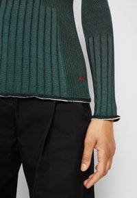 Personne portant un pantalon noir et un haut à manches longues côtelé vert foncé avec des rayures verticales et une bordure blanche aux poignets et à l'ourlet.