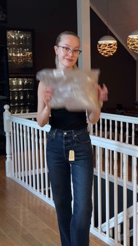 Transparent plastförpackning hålls av en person med texturerad yta. Personen bär mörkblå jeans och svart topp, står på ett trägolv.