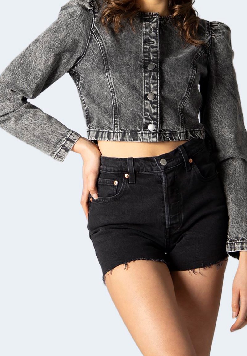 Chaqueta de mezclilla gris recortada con mangas abullonadas y botones plateados, combinada con pantalones cortos de mezclilla negros de tiro alto con dobladillos deshilachados.