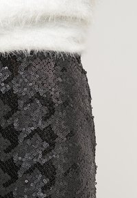 Svart paljettkjol med en glänsande, texturerad yta. Har en figurnära design och är delvis täckt av en luftig vit tröja.