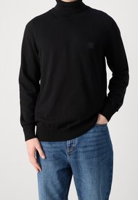 Pull à col roulé noir avec poignets et col côtelés, arborant un petit écusson noir avec logo sur la poitrine. Associé à un jean en denim bleu.