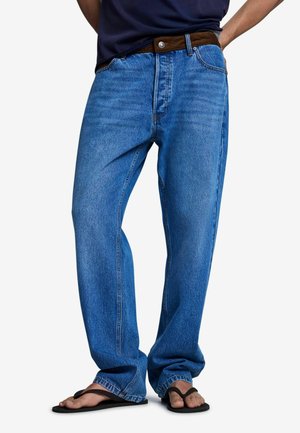 Personne portant un jean bleu ample avec une ceinture marron et des tongs noires, debout avec une main dans la poche.
