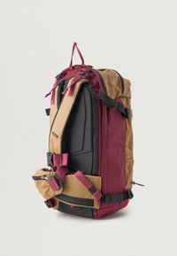A mochila possui um topo bordô, lados com padrão de grelha bege, um painel preto, alças ajustáveis e múltiplas fivelas externas para um fecho seguro.