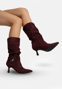 Bottes basses en daim bordeaux au design ample, bout pointu et talon stiletto de 10 cm, avec un motif décoratif cousu et un accent de quincaillerie.