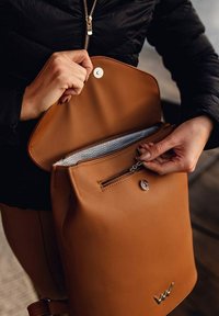 Hnědý faux kůže batoh s uzavíráním na zip, předním zapínáním na patentku a interiérem s chevronovým vzorem; vybaven stříbrným kováním.