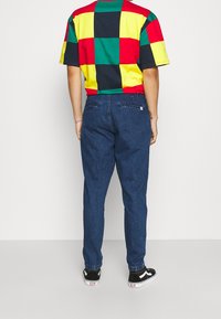 Camisa de manga corta a cuadros multicolor con cuadrados rojos, amarillos, verdes y negros, combinada con pantalones de mezclilla azul oscuro y zapatillas negras.