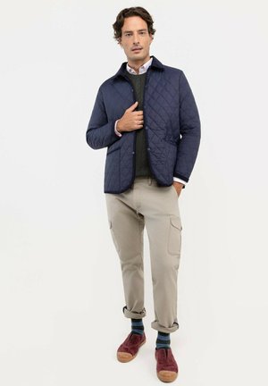 Hombre con chaqueta acolchada azul marino, suéter oscuro, pantalones cargo beige, calcetines a rayas y zapatos casuales rojos, de pie frente a un fondo liso.