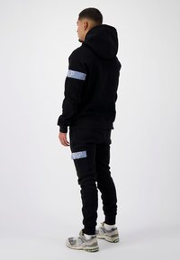 Zwarte hoodie en bijpassende joggers van een zachte stof, met blauwe accenten op de armen en zakken. Model draagt multicolore sneakers.