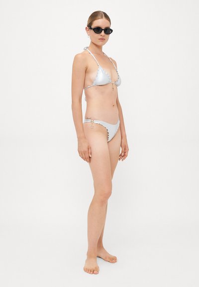 Sieviņš ar sudrabotu metālisku bikini, kas rotāta ar zvaigžņu piekariņiem, melnās ovālas saulesbrillēs, stāvot basām kājām pret vienkāršu baltu fonu.