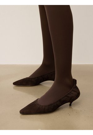 Pumps de ante marrón con punta afilada y un suave borde fruncido, con un tacón pequeño, llevados con medias marrón oscuro sobre una superficie de concreto.