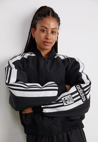 Svart bomberjakke med hvite striper langs ermene og ribbestrikkede mansjetter, med Adidas-logo på venstre erme. Glatt, polstret tekstur.