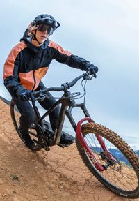 Horská kola v černé a červené barvě, s pevným rámem a silnými, texturovanými pneumatikami. Jezdec má na sobě černou a oranžovou bundu, helmu a sluneční brýle.