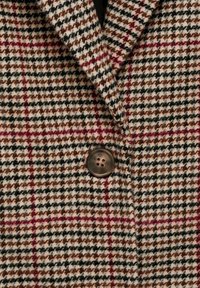 Plašč iz volnenega mešanice s houndstooth vzorcem v bež, rjavi in burgundski barvi, z odrezanimi klapnicami in enim rjavim gumbom.