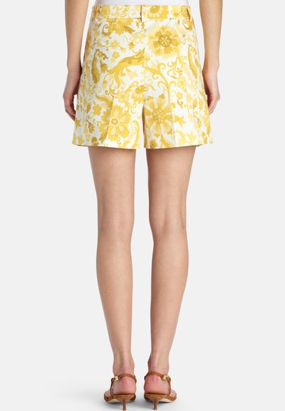 Femme portant un short blanc taille haute à imprimé floral jaune, debout pieds nus sur un fond blanc uni.