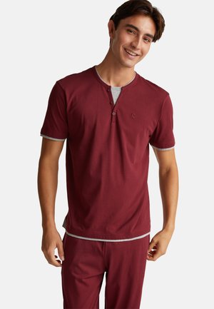 Joven sonriente que lleva una camisa Henley de manga corta color burdeos con ribetes grises y pantalones burdeos a juego, de pie contra un fondo blanco.