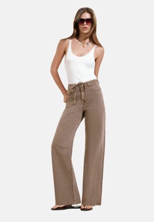 Frau mit großen Sonnenbrillen, weißem ärmellosem Oberteil, braune Hose mit hohem Bund und weitem Bein, schwarzen Sandalen und einer bunten Perlenkette.