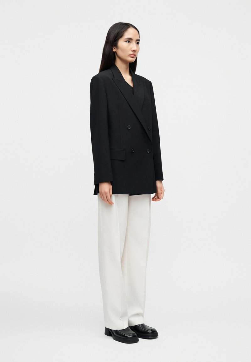 Blazer noir croisé à gros boutons, associé à un pantalon blanc à jambes larges et des mocassins noirs à plateforme. Design élégant et structuré.