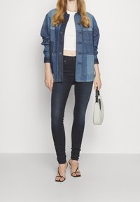 Chaqueta de denim con tonos azules variados, grandes bolsillos frontales y cierres de botón negros; combinada con jeans ajustados negros y tacones negros de tiras.