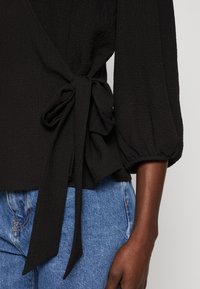 Sort tekstureret bluse med wrapsdesign, der har et bånd i taljen og ballonærmer. Sammen med blå denim.