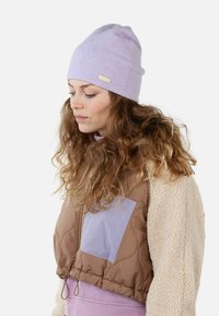 Lilac beanie with a folded brim and a small branded tag. Worn with a brown quilted vest featuring a light purple pocket.