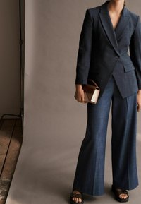 Žena v tmavě modrém dobře padnoucím sakě a širokých kalhotách, která drží malou hnědou kabelku a má na sobě černé sandály na otevřené špičce.