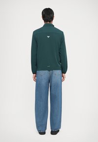 Σκούρο πράσινο πουκάμισο με μακριά μανίκια και γιακά, με λογότυπο στο πίσω μέρος. Συνδυάζεται με φαρδιά μπλε τζιν και μαύρα παπούτσια.