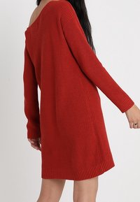 Rotes gestricktes Kleid mit lockerem Schnitt, schulterfreiem Design, langen Ärmeln, gerippter Textur und Saum, der knapp über dem Oberschenkel endet.