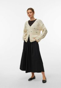 Kremast pleten pulover z vezenjem cvetja, sprednjimi žepi in gumbi, kombiniran z dolgo črno krilo in črnimi ravnimi čevlji.