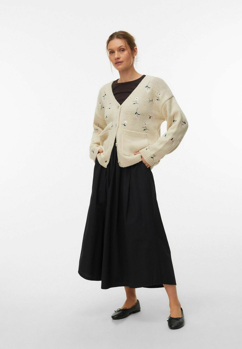 Kremast pleten pulover z vezenjem cvetja, sprednjimi žepi in gumbi, kombiniran z dolgo črno krilo in črnimi ravnimi čevlji.