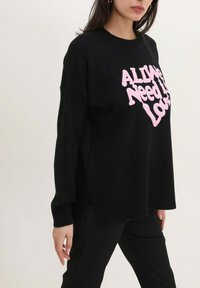 Pull noir surdimensionné avec ourlet côtelé présentant un texte rose vif "Tout ce dont nous avons besoin, c'est d'amour". Tissu en maille douce, manches longues, fentes sur les côtés.