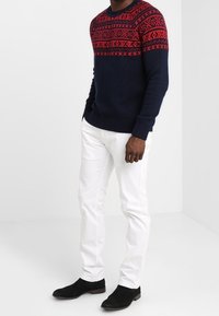 Marineblauwe gebreide trui met rode geometrische patronen op de schouderpartij, gecombineerd met witte broek en zwarte enkellaarsjes. Eenvoudig, gestructureerd ontwerp.