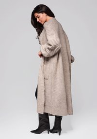 Langer beiger Strickcardigan mit lockerer Passform, strukturiertem Muster, seitlichen Taschen, gerippten Bündchen und leicht verlängertem Saum, kombiniert mit schwarzen knielangen Stiefeln.