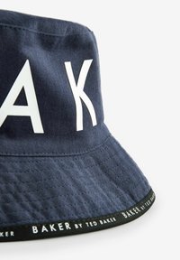 Pălărie de tip bucket de culoare albastră marină, fabricată din bumbac, cu litere albe mari, finisaj texturat și o bandă neagră cu logo-ul brandului în jurul marginii.