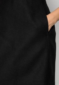 Vêtement en laine noire avec une poche latérale, présentant une texture lisse et une silhouette ajustée.