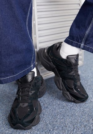 Czarne masywne sneakersy noszone z białymi skarpetkami i ciemnoniebieskimi dżinsami, ustawione na niebieskim dywanie blisko metalowej kratki wentylacyjnej.