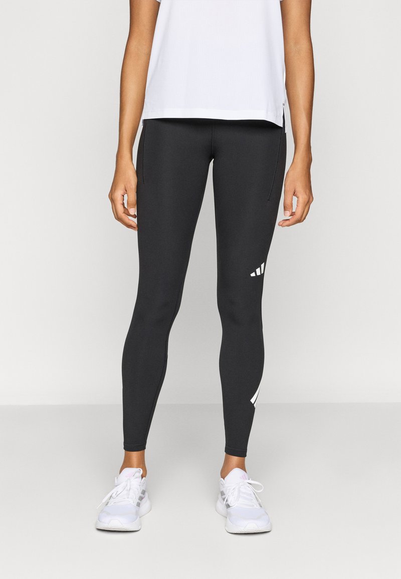 Schwarze Leggings mit glatter Textur, weißen Logoakzenten, figurbetontem Schnitt und hohem Bund. Kombiniert mit einem weißen Oberteil und Sportschuhen.