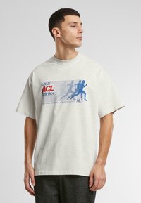 Tricou supradimensionat, de culoare gri deschis, confecționat din bumbac, cu un design grafic albastru și roșu, ce include text și siluete de alergători. Mâneci scurte.