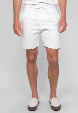 Homme portant un short blanc à cordon, des chaussures blanches sans lacets et une chemise blanche, debout devant un fond uni clair.