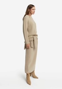 Beiges Strickkleid mit langen Ärmeln und gebundenem Taillengürtel, kombiniert mit beigen Knöchelstiefeln. Der Stoff weist ein strukturiertes, geripptes Muster auf.
